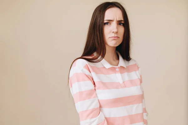 The frowning girl clenches her brows and lips, puffs her nostrils angrily, and puffs aggressively. Beautiful young woman dressed in a striped cardigan stands on a beige background in the studio