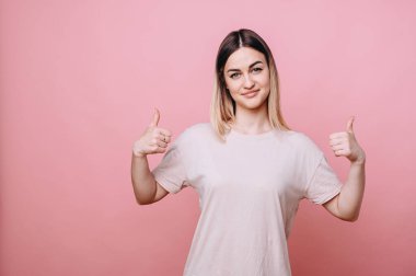 Sıradan giyinmiş bir kız, iki eliyle baş parmağını kaldırıyor ve izole bir arka planda nazikçe gülümsüyor..