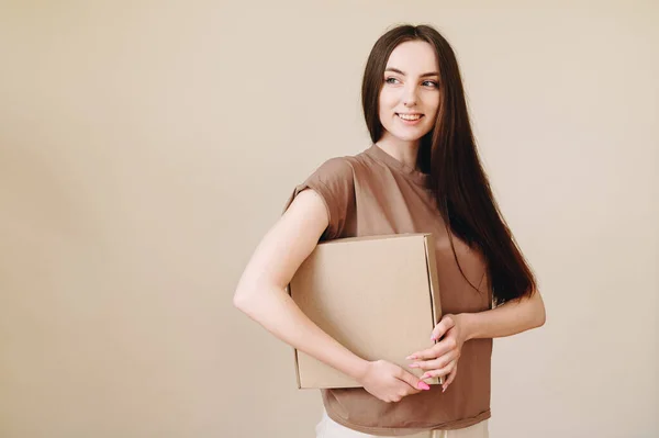 A young well-groomed girl holds a cardboard box on hand and looks at the camera on a beige background. Concept to receive mail.