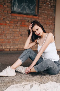 Kısa saçlı genç bir kız başı bir yana eğik şekilde oturur, elini tutar ve kameraya bakar. Siyah kot giyen bir kızın dikey fotoğrafı..