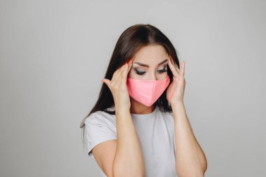 Beyaz tişörtlü esmer bir kadın pembe koruyucu maskeli beyaz bir arka planda elini şakaklarına uzatıp aşağı bakıyor. Hastalık, grip konsepti.