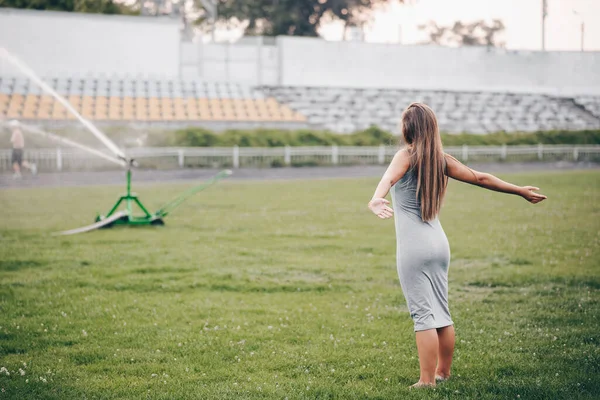 一个穿着灰色衣服的女孩站在那里 双手分开 等待着 草坪洒水器在体育场里喷水 夏天的傍晚在草地上洗澡 — 图库照片