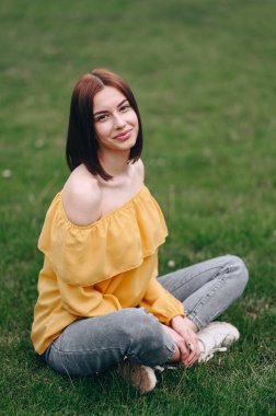 Kısa saçlı bir öğrenci yeşil çimlerin üzerinde parlak sarı bir kazakla bacak bacak üstüne atarak oturur. dikey resim