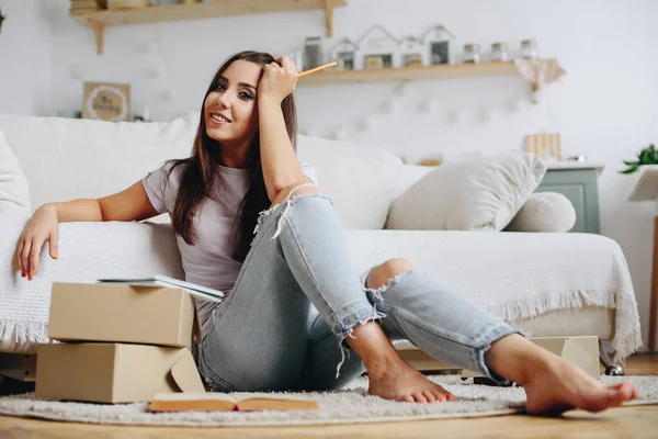 A pretty brunette girl with a smile sits in front of a sofa looking at the camera holding a pencil in her hands, among cardboard boxes. Online delivery, freelance manager.