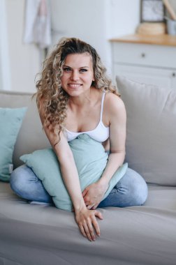 Yumuşak koltukta oturan, yastığa sarılan ve kameraya neşeyle bakan kıvırcık sarışının dikey portresi.