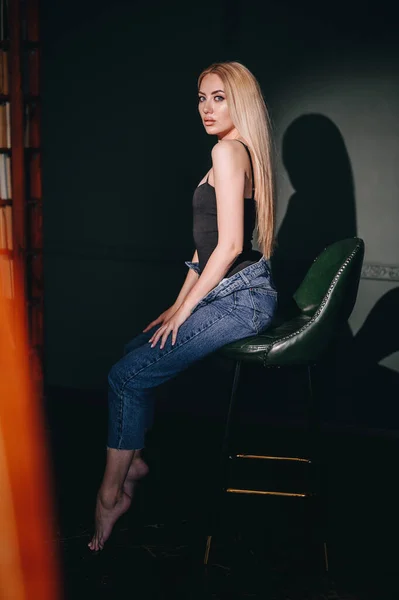 ハードスタジオライト付きの緑の壁に対して椅子に座っている女性の垂直写真 長いブロンドの髪のジーンズの女の子 手を触れずに — ストック写真
