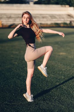 Kız stadyumdaki spor kıyafetlerinde dizlerini kaldırıyor. Saçları dökülmüş dikey fotoğrafçı kızlar yeşil çimlerde egzersiz yaparlar. Boşluğu Kopyala.