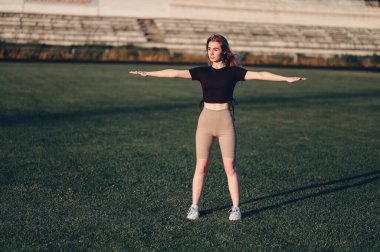 Siyah bluzlu kız kollarını yana doğru açmış egzersiz yapıyor. Full Height Woman Green Lawn 'daki stadyumda duruyor. Koşmadan önce ısınıyor.