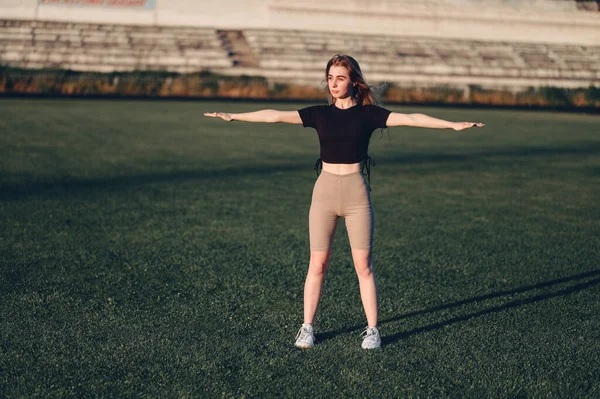 Siyah bluzlu kız kollarını yana doğru açmış egzersiz yapıyor. Full Height Woman Green Lawn 'daki stadyumda duruyor. Koşmadan önce ısınıyor.