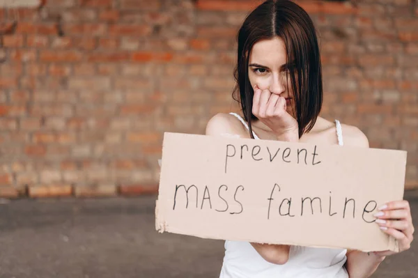 Brunette woman with cardboard hands to stop mass famine. Cover your mouth with your fist and look from under your forehead.