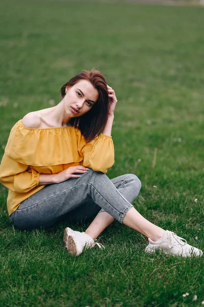 Młoda Kobieta Żółtej Bluzce Dżinsach Siedzi Trawie Wygląda Zainteresowaniem Pionowe — Zdjęcie stockowe