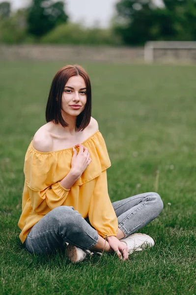 Young Woman in Yellow Blouse and Jeans Sits in Lotus Position on Grass in the Park. Vertical Photo of Brunette Woman with Short Hairstyle Outdoors, Sat Down to Relax After Working Day.