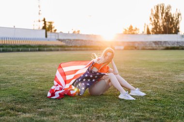 Sarışın, Yeşil Çimenler Üzerinde Amerikan Bayrağına Sarılmış Oturuyor. Batan Güneş Işıkları Stadyumundaki Genç Kadın. Bayrak rüzgarda sallanıyor. Bağımsızlık Günü Kavramı