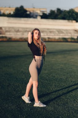 Genç bir kadın yeşil çimlerin üzerinde güneş ışınlarıyla egzersiz yapıyor. Spor Giyim ve Spor Ayakkabılı Bir Kızın Dikey Portresi. Bir yaz akşamında vücudunu boşalt..