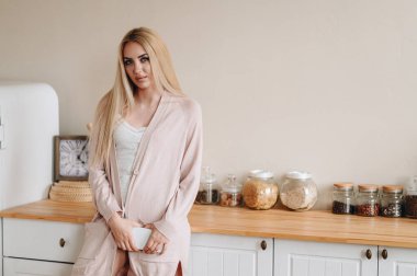 Sarı saçlı bir kadın mutfağın arka planında duruyor. Kız elinde bir telefon tutuyordu. Kadın, kocasını evde elbiseleriyle bekliyor..