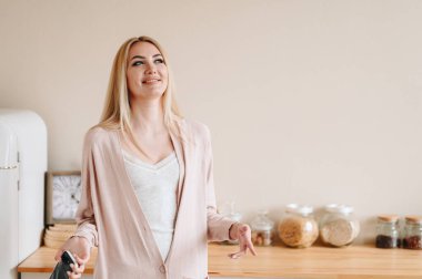 Ellerinde telefonu olan sıradan kıyafetlerle sarışın bir kız kendi kendine gülüyor. Hatalarına gülün. Ev samimi iç mekan. Mutfak aletlerinin arka planındaki kız