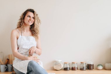 Önlükteki Genç Eş Mutfakta Masada Oturuyor ve Neşeli Uzakta Bakıyor. Kadın yemek blogunu yönetiyor. Konforlu Ev İçi.