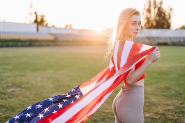 Atletik Zayıf Kadın güneşe karşı duruyor ve Amerikan bayrağını omuzlarında tutuyor. Yakın plan portre. Doğada Yaz Akşamı.