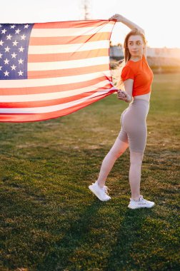 Kırmızı Spor Üstündeki Dikey Fotoğrafçı Kadın ve Uzun Boylu Amerikan Bayrağı Dalgalanıyor. Yeşil Çimenlikteki Spor Kızı bayrağın yanında duruyor. Amerika 'da Bağımsızlık Günü kavramı