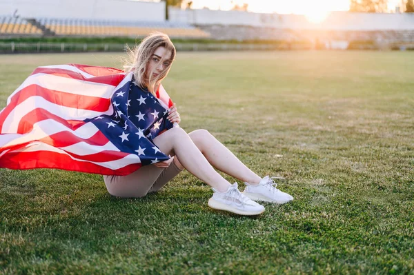 Sıcak bir yaz akşamında, Atletik bir kadın güneşe karşı oturur ve omuzlarında bir Amerikan bayrağı taşır. Şirin Gülümseyen Kızın Yakın Çekim Portresi.