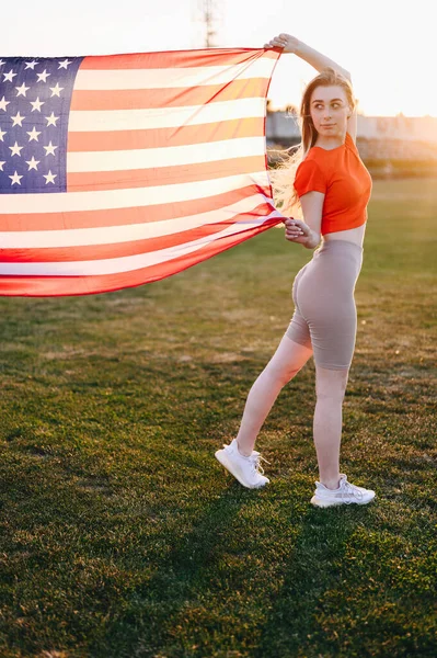 Vertical Photo Woman in Red Sports Top and Tights Holding American Flag Waving in Wind in Full Length. Sports Girl on Green Grass stands with Flag. Independence Day concept in America