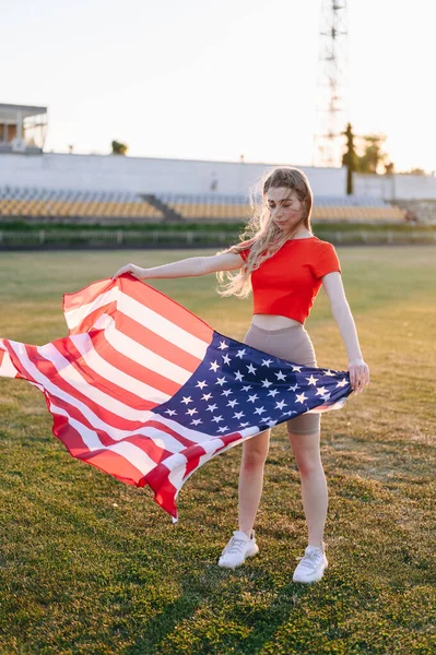 Vertical Photo Woman in Sports Top and Tights Holding American Flag Waving in the Wind. Sports Girl on Green Grass stands with a Flag.