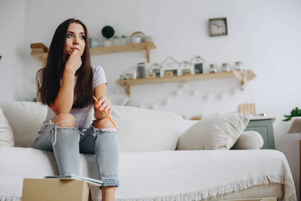 A young woman in torn jeans sits pensively on a sofa. Bright interior in natural light. Pencil in hand. Concept Unemployed.