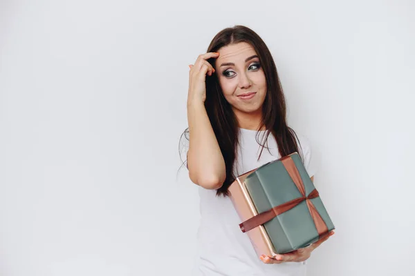Brunette Woman Holds Gifts in Her Hands. Think about what to give for Holiday. Girl Scratches her Forehead and Grimaces. Girl on White Background.