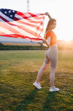 Amerikan Bayraklı Kız 'ın Tam Uzun Boylu Dikey Fotoğrafı. İki eli de rüzgarda dalgalanan bayrak tutan kızlar. Anma Günü Spor Kızı. Altın Saat 'te gün batımı.