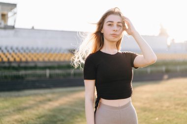 Siyah Üstlü Genç Kadın 'ın portresi, Saçını Rüzgardan Ayarlıyor. Kız spor sahasının arka planında duruyor. Yan Günbatımı Işığı.