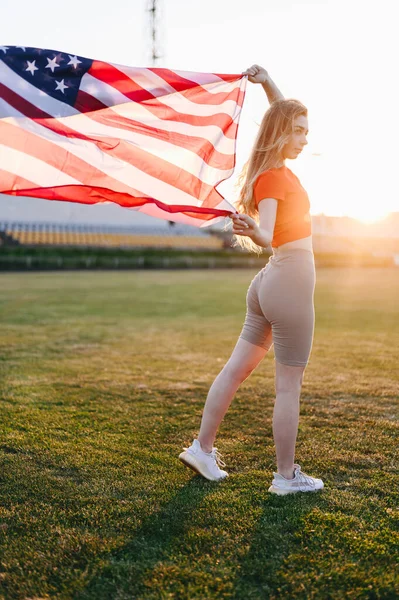 Full Length Vertical Photo of Girl with an American Flag. Girl Holds with both hands flag fluttering in wind. Memorial Day Sports Girl. Sunset at Golden Hour.
