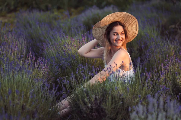 Beautiful Happy Woman in Lavender Field at Golden Hour at Sunset. Summer Evening in Lavender Field. Young Woman with Sincere Smile sits in Hat and Looks at Sunset.