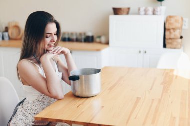 Önlüklü genç kadın ahşap bir masada oturur ve bir tencereye bakar. Uzayı kopyala Ne pişireceğini düşün.