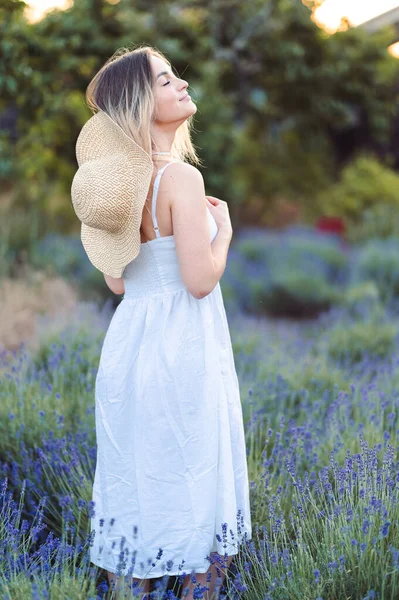 Beautiful Young Woman in Summer Dress and Straw Hat Inhales Fresh Fragrance of Lavender. Girl Enjoys Summer in Lavender Field