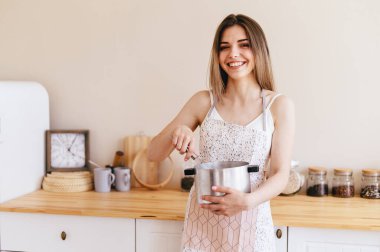Happy Young Woman Holds Saucepan in Her Hands and Stirs with Whisk. Girl in Apron prepares delicious dish. Light Cozy Kitchen in Background