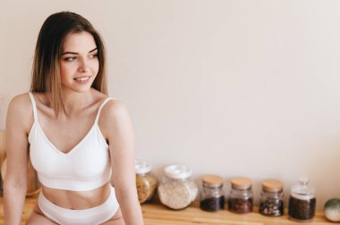 Seksi genç kadın mutfak masasında oturur öne eğilir ve pencereden dışarı bakar. Bej Mutfak 'ta beyaz iç çamaşırlı bir kız. Uzayı kopyala Güne harika bir başlangıç.