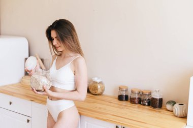 Seksi Genç Kadın Mutfağın Arkaplanına Karşı Duruyor ve Elinde Yulaf Ezmesi Kavanozu Tutuyor. Bej Mutfaktaki Beyaz İç Çamaşırlı Kız. Evdeki rahat iç mekan. Güne Harika Başlama.