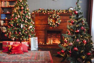 Wooden Fireplace is Decorated with Christmas Decor. Christmas Tree with Red Toys in Front and Background. New Year Cozy Home Interior.