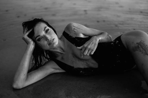 Young Wet Woman in One-Piece Swimsuit lies on Shore on her Side Holding her Head. Woman Smeared with Therapeutic Mud. Black and White Photography