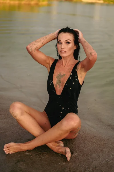 Young Woman in Black Bathing Suit Sits Cross-Legged on Shore. Woman Straightens Wet Hair. To be Smeared with Therapeutic Mud.