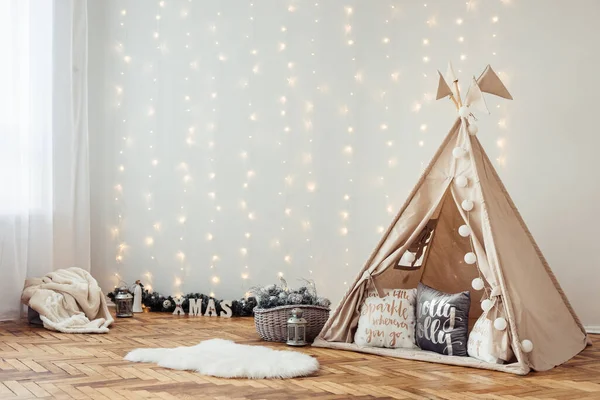 Children's New Year's Photo Zone with Wigwam Against Background of Luminous Electric Garland on Wall. Parquet Floor in Interior.