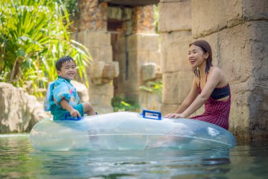 Asyalı çocukla annesi yazın yüzme havuzunda mayo giyip su parkında oynuyorlar.