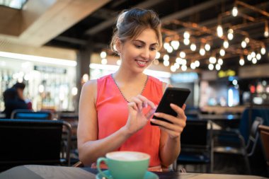 Genç bir kadın kahve içerken akıllı telefonunu kullanıyor.