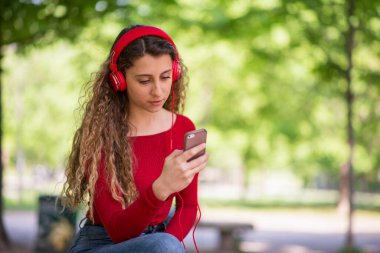 Kırmızı elbiseli genç parkta telefondan müzik dinliyor.