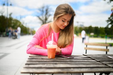 Mutlu genç kadın akıllı telefonunu kullanırken açık havada smoothie içiyor.