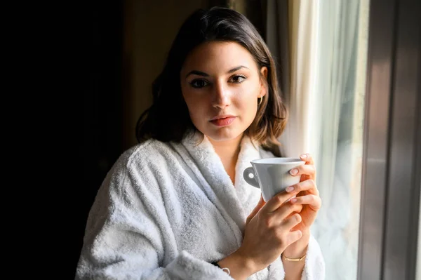 バスローブを着てホテルの部屋の窓の近くでコーヒーを飲む若い女性 — ストック写真