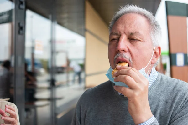 男食べる彼のファーストフードランチハンバーガー中にマスクで彼のあご コロナウイルスの概念 — ストック写真