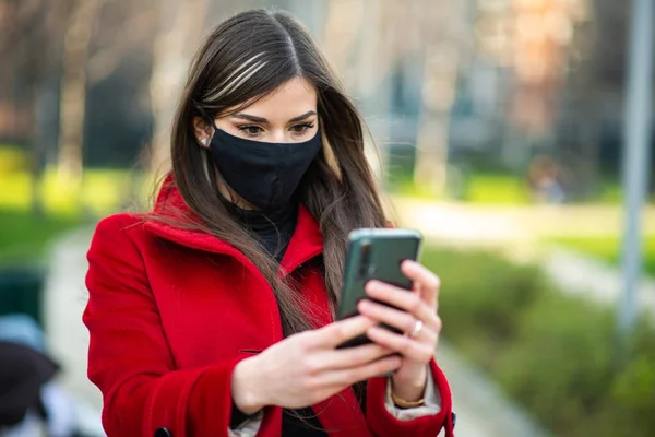 コロナウイルスの緊急時にスマートフォンを使用している間 市内公園を歩く仮面の女性 — ストック写真