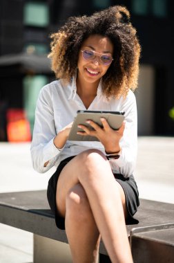 Gülümseyen Afro-Amerikan iş kadını dijital tablet kullanarak bankta oturuyor.
