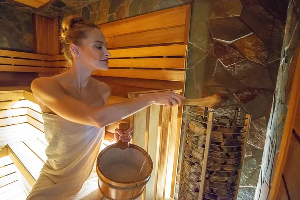 Woman in a sauna putting water on hot rocks to produce more steam